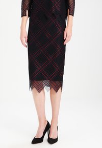 Svart spetsklänning med röda och marinblå geometriska mönster, figursydd silhuett, vågig nederkant och trekvartslånga ärmar. Kombinerad med svarta klackar.