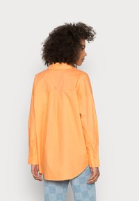 Camisa laranja de botão, com mangas longas e um corte descontraído. Apresenta uma pala nas costas e um pequeno detalhe de logótipo. Usada com calças xadrez azuis.