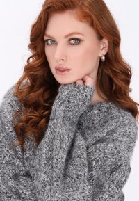 Grau gestrickter Pullover mit lockerem Schnitt und gerippten Bündchen. Modell mit langen, welligen roten Haaren präsentiert einen hohen Kragen und ein weiches, strukturiertes Material.