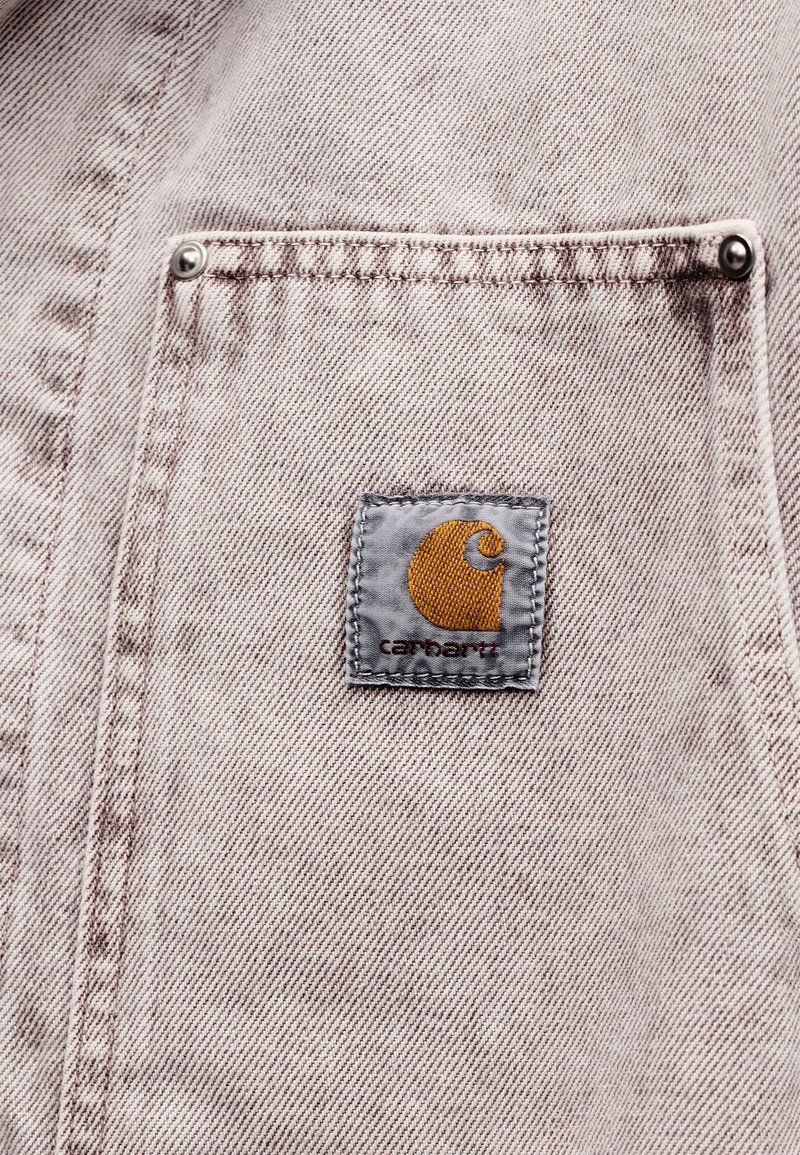 Tissu en denim beige avec une petite poche avant comportant un patch carré du logo Carhartt cousu près du centre.