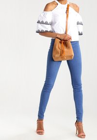 Biała bluzka bez ramiączek z czarnym haftem, niebieskie dopasowane dżinsy i beżowa zamszowa torba w kształcie wiadra. Różowe sandały na obcasie dopełniają outfit.