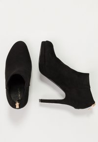 Černé semišové kotníkové boty s špičatou špičkou, vysokým stiletto podpatkem a bočním zipem se zlatým akcentem. Hladká textura a minimalistický design.