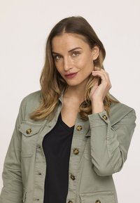 Veste vert pâle à boutons, avec deux poches poitrine et des boutons en écaille de tortue. Portée par-dessus un t-shirt noir à col en V. Texture lisse, design décontracté.