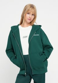 Felpa verde con cappuccio e zip frontale, tasche a marsupio e logo bianco sul petto. Indossata sopra una t-shirt bianca e pantaloni verdi.