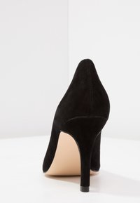 Chaussure à talon haut en daim noir avec un bout pointu et un talon fin, présentant une texture lisse et une semelle intérieure de couleur neutre.