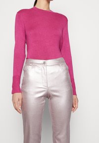 Sittande rosa stickad tröja i kombination med glänsande silvriga metallbyxor med knappstängning och sidofickor. Slät textur överlag.
