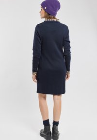 Marineblauwe jurk met lange mouwen en een kraag en manchetten van ruitpatroon, afgewerkt met stevige zwarte enkellaarsjes en een paarse gebreide beanie.