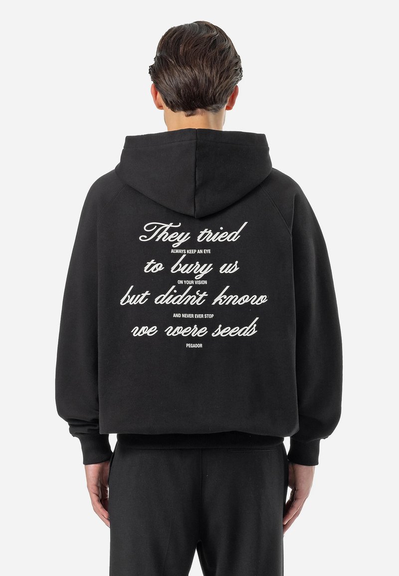 Black hoodie with a spacious hood, featuring white text on the back that includes various phrases and a brand name, made from soft fabric.