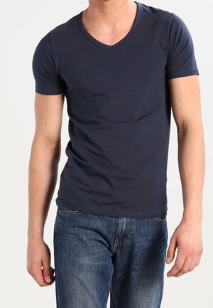 Mężczyzna ubrany w dopasowaną granatową koszulkę z krótkim rękawem i dekoltem w serek oraz niebieskie dżinsy na jednolitym tle.