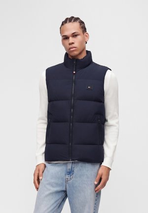 Ung mand med flettet hår iført en sort dynevest over en hvid sweater og lyseblå jeans mod en ensfarvet baggrund.