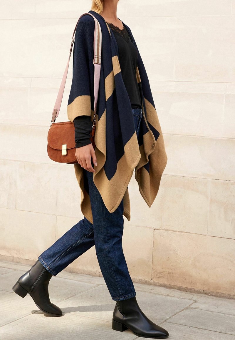 Femme marchant, portant un poncho ouvert bleu marine et beige, un haut avec bordure en dentelle noire, un jean foncé, des bottines noires, et portant un sac bandoulière marron.