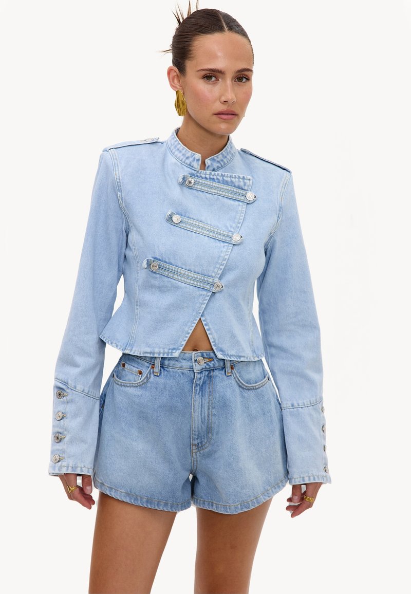 Femme portant une veste en jean bleu clair avec des bandes de boutons asymétriques et un short en jean assorti, debout devant un fond uni.