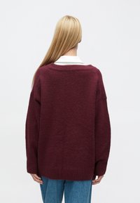 Vínovočervený sveter s textúrou, voľného strihu, s rebrovanými manžetami a lemom, vrstvený cez bielu košeľu s golierom. Materiál pripomínajúci vlnu; pohľad zozadu.