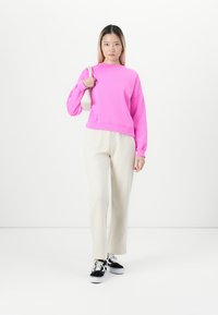 Young woman stands wearing bright pink sweatshirt, cream pants, black sneakers, and carrying small white shoulder bag.