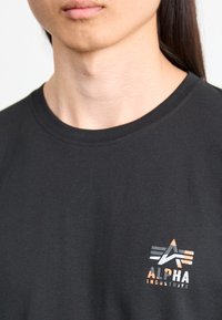 Tricou din bumbac negru, cu un mic logo grafic portocaliu și alb, cu un design unghiular, amplasat în zona stângă a pieptului. Mânici scurte, gât rotund.