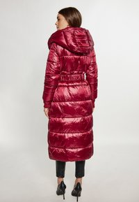 Palton puffer lung burgund cu glugă și talie strânsă, având o textură lucioasă și un design matlasat orizontal. Pantaloni negri și tocuri vizibile.