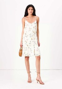 Rochie midi florală în crem, cu bretele subțiri, tiv ondulat și talie ajustabilă. Asortată cu o geantă maro și sandale cu curele.