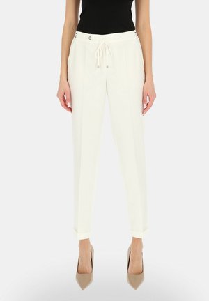 Kobieta nosząca białe, zwężane spodnie z wysokim stanem i troczkiem w talii, czarny top bez rękawów oraz beżowe szpiczaste szpilki.