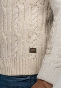 Pull en maille torsadée de couleur crème avec ourlet et poignets côtelés, porté avec un jean denim bleu, avec une petite étiquette de marque en cuir marron.