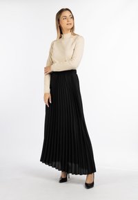 Lang sort plisseret nederdel med elastisk talje, parret med en lys beige langærmet top, begge designet til en tætsiddende pasform. Sorte højhælede sko fuldender outfittet.