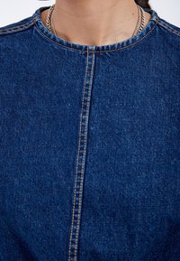 Top de mezclilla con cuello redondo, que presenta costuras contrastantes en claro a lo largo de la costura central y el cuello. Tela azul oscuro, textura suave.