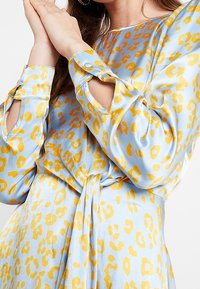 Vaaleansininen satiinimekko, jossa on keltainen leopardikuosi. Mekossa on pitkähihaiset hihat, solmittava vyötärö ja napit hihansuissa.