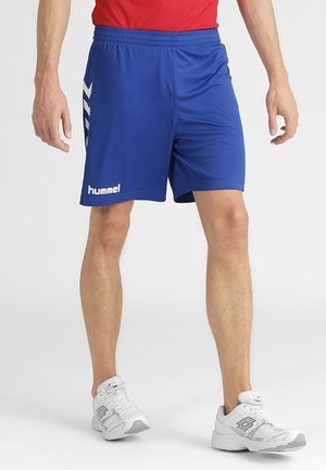 Mand iført blå Hummel sports-shorts, rød skjorte og hvide atletiksko stående mod en ensfarvet baggrund.