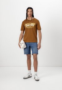 Camiseta de algodón marrón con texto gráfico en blanco y amarillo, combinada con pantalones cortos de mezclilla azules y zapatillas blancas con acentos negros.