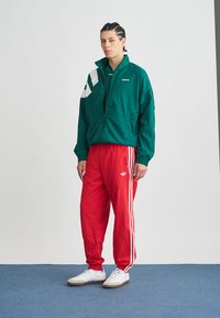 Green jacket with white accents, red pants with white stripes, and white sneakers. Smooth fabric, relaxed fit, and zipper details.