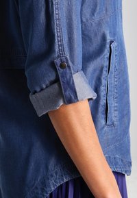 Giacca di jeans con maniche arrotolate, con un polsino interno blu a contrasto e un dettaglio con bottone a pressione. Tasca laterale visibile nell'area del torace.