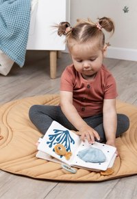 Zacht stoffen boek met kleurrijke illustraties, waaronder een vis en een schelp, in blauwtinten, geel en wit. Kind zit op een ronde, beige mat.
