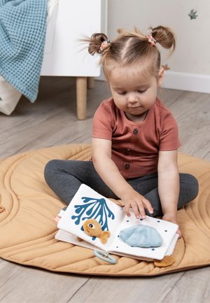 Pehmeä kangaskirja, jossa värikkäitä kuvituksia, kuten kala ja simpukka, ja sinisen, keltaisen sekä valkoisen sävyjä. Lapsi istuu pyöreällä, beigen värisellä matolla.