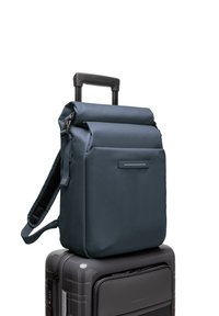 Sac à dos à rabat bleu foncé en tissu texturé, avec une poche avant et une bandoulière réglable, posé sur une valise grise élégante.