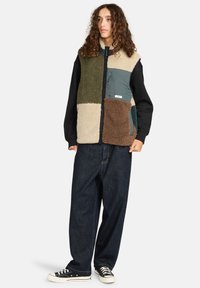 Kamizelka z polarowego materiału w kolorach beżowym, zielonym i brązowym, z zapięciem na zamek błyskawiczny, kieszeniami, noszona na czarnej bluzie z długim rękawem i luźnych dżinsach.