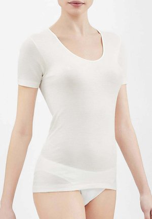 Donna che indossa una camicia bianca aderente a maniche corte e biancheria intima bianca su uno sfondo bianco semplice.