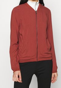 Veste bomber rouge avec fermeture éclair, poignets côtelés et poches latérales. Confectionnée en tissu léger, elle présente une texture lisse et un design minimaliste.