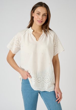 Blouse blanche brodée avec des manches et un ourlet en forme de scallop, ornée de motifs floraux découpés. Assortie avec un jean bleu clair.