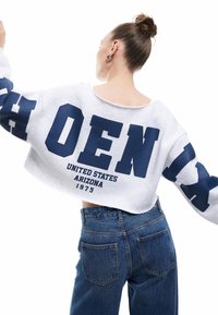 Sudadera blanca recortada con texto en navy "PHOENIX ESTADOS UNIDOS ARIZONA 1975" y grandes detalles en navy en las mangas. Combinada con jeans azules.