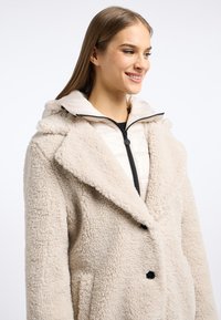 Lichtbeige schapenvachtjas met een pluche textuur, grote kraag, knoopsluiting en zwarte accenten. Beschikt over een verwijderbare crèmekleurige binnenjas met rits.