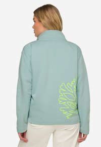 Světle modrá fleecová bunda s vysokým l�ímcem, která má na zadní straně velký neonově zelený grafický design. Měkká textura a volný střih.