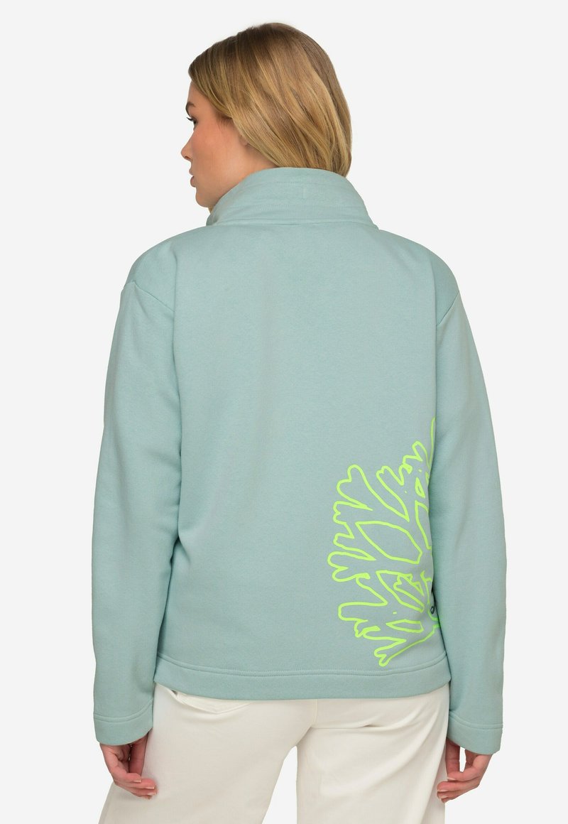 Světle modrá fleecová bunda s vysokým límcem, která má na zadní straně velký neonově zelený grafický design. Měkká textura a volný střih.
