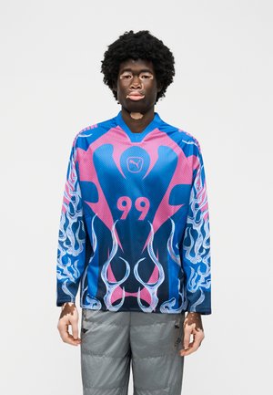 Osoba s vitiligem, oblečená v modro-růžovém dresu s plamenovým vzorem a číslem 99, s šedými kalhotami, stojící před jednobarevným pozadím.