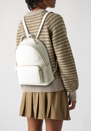 Zaino in pelle bianca morbida con design arrotondato. Presenta una tasca anteriore con zip e hardware color oro. Indossato sopra un maglione a coste e una gonna.