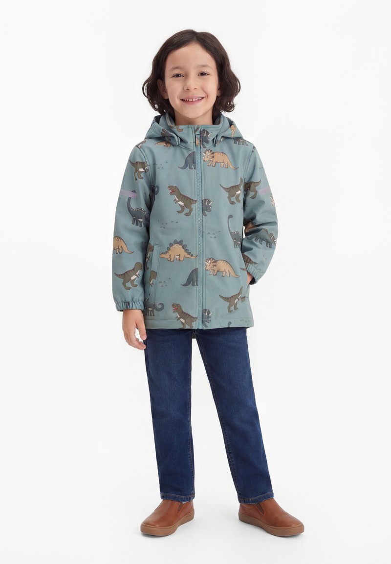 Παιδί που στέκεται και χαμογελά, φορώντας μπλε ζακέτα με κουκούλα και σχέδιο δεινόσαυρο, σκούρο μπλε τζιν και καφέ παπούτσια τύπου slip-on.