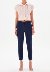 Cropped top a righe in rosso e bianco, abbinato a pantaloni slim-fit blu scuro con dettagli in oro sui bottoni. Il modello indossa tacchi bianchi con cinturini.
