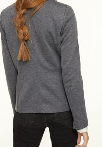 Harmaa, teksturoidulla pinnalla varustettu bleiseri, jossa istuva leikkaus ja hillitty kuviointi. Henkilöllä on päällään tummat farkut ja valkoinen paita.