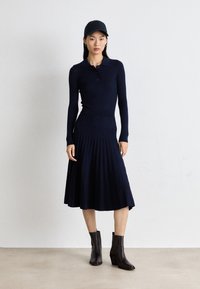Navy ribbed knit dress with a polo collar, long sleeves, and a pleated skirt. Paired with dark ankle boots and a black cap.