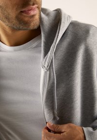 Homme avec une légère barbe portant une chemise blanche, tirant la fermeture éclair d'un sweat à capuche gris d'une main.