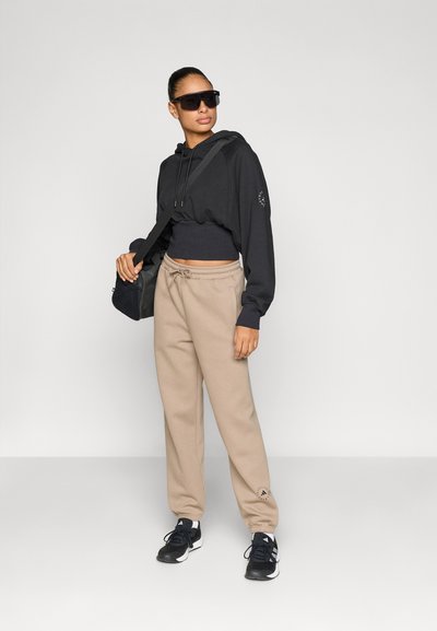 Μαύρο κολαριστό φούτερ με κουκούλα, μπεζ φόρμα και μαύρα αθλητικά παπούτσια. Το φούτερ έχει κορδόνι και λογότυπο στο μανίκι· το παντελόνι διαθέτει ελαστικά μανσέτα.