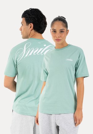 Mand og kvinde iført lysegrønne T-shirts med teksten "Smilodon"; manden vender ryggen til, kvinden vender frem, begge iført lysegrå bukser.